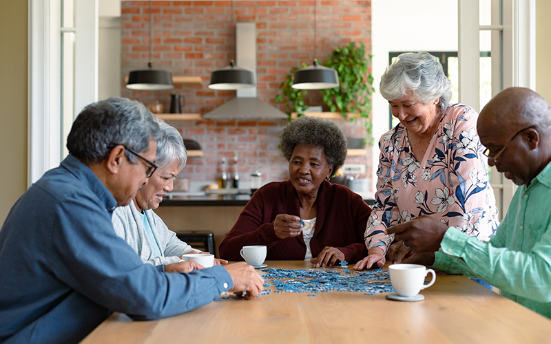 Jogos como quebra-cabeça ajudam a exercitar a memória. Cinco pessoas com xícaras de chá branca e peças de quebra-cabeça sobre uma mesa de madeira.
